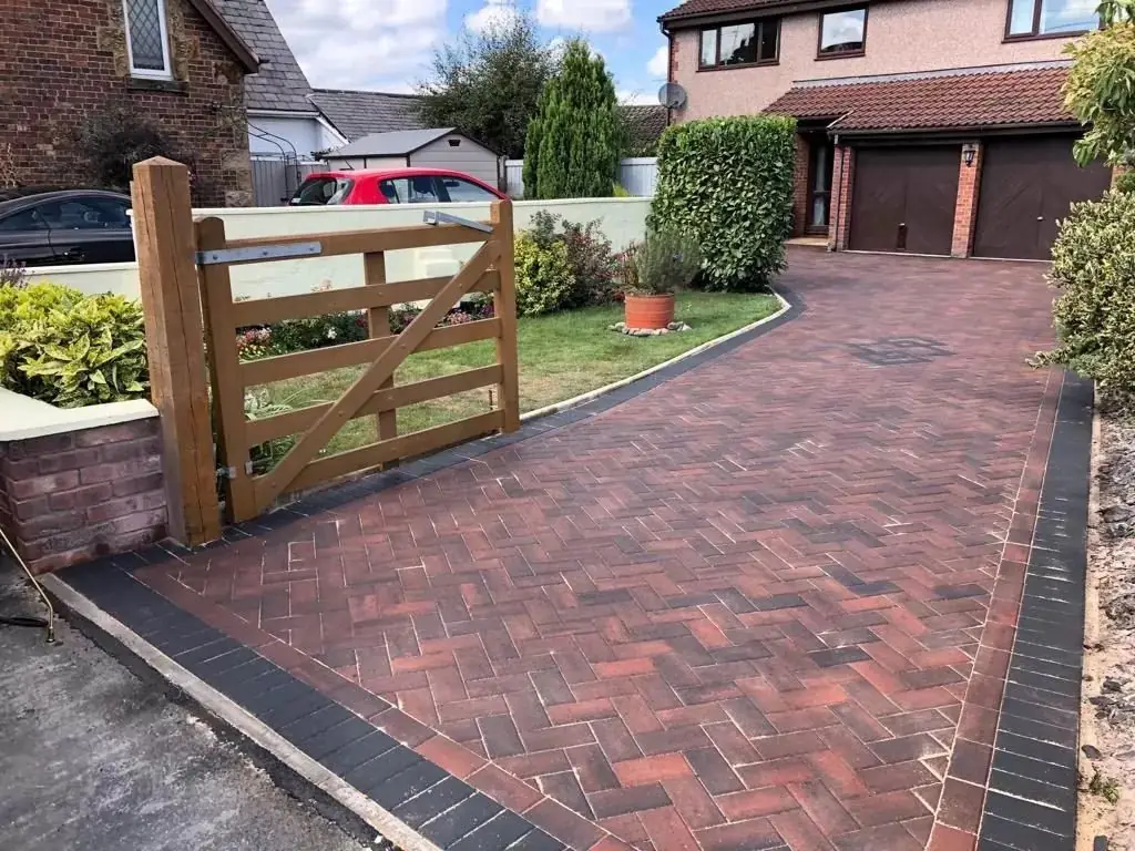 Red brindle block paved driveway laid in herringbone with a charcoal border, running past a timber gate up to the garage